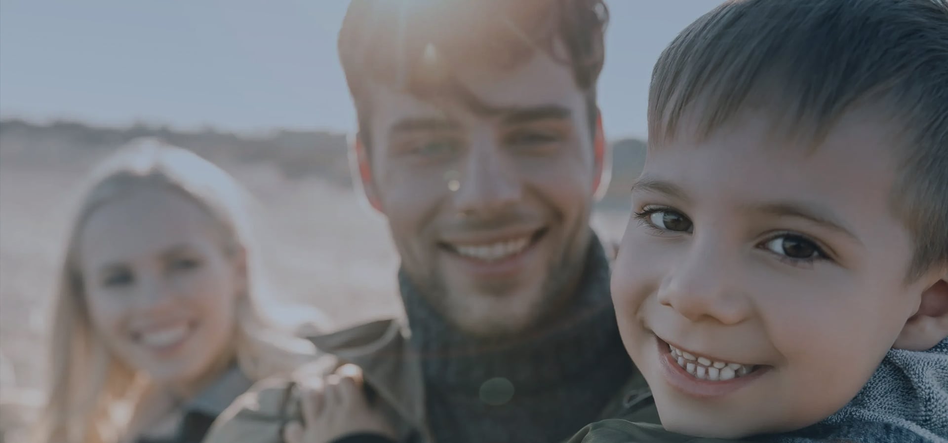 A father holding his smiling young son outdoors, with the child's mother beside them.