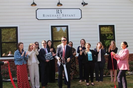 A wide group shot of the Rinehart Bryant team and guests posing with a red ribbon and oversized scissors outside the Spotsylvania family law office.