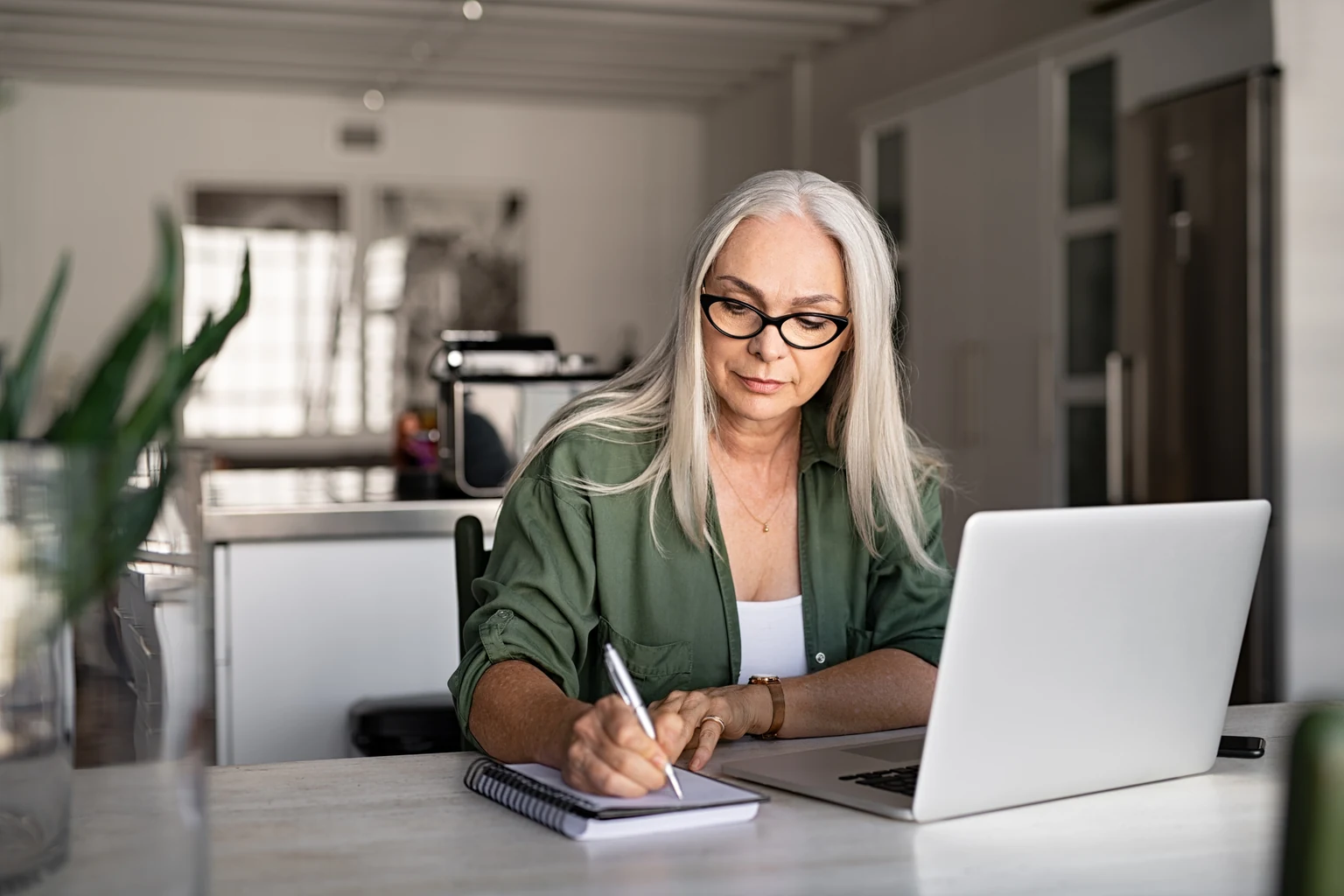 A woman with white hair sits at her kitchen counter writing in a notebook beside an open laptop